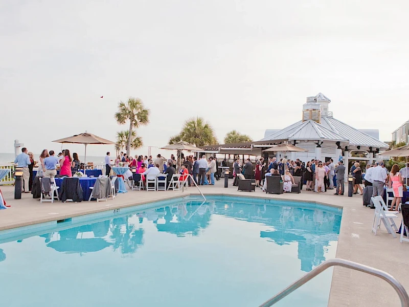 A poolside party or wedding reception with guests mingling by tables and a gazebo, palm trees, and a bright, sunny outdoor setting.