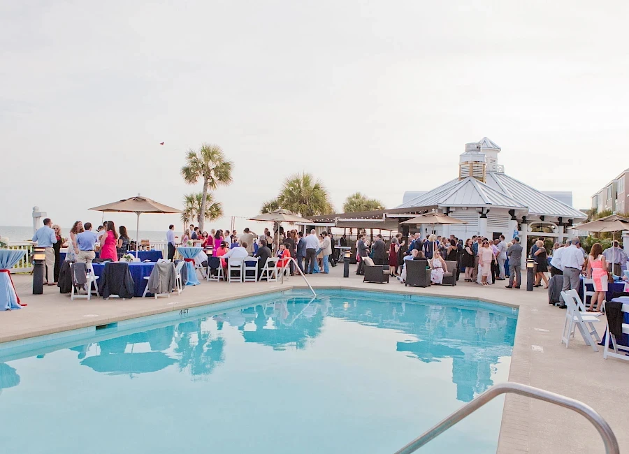 A poolside party or wedding reception with guests mingling by tables and a gazebo, palm trees, and a bright, sunny outdoor setting.