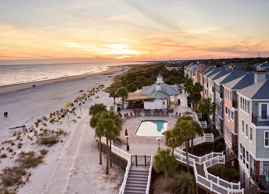 A beachside condo complex with a pool, palm trees, and a sandy shoreline at sunset, overlooking the ocean along a narrow coastal town.