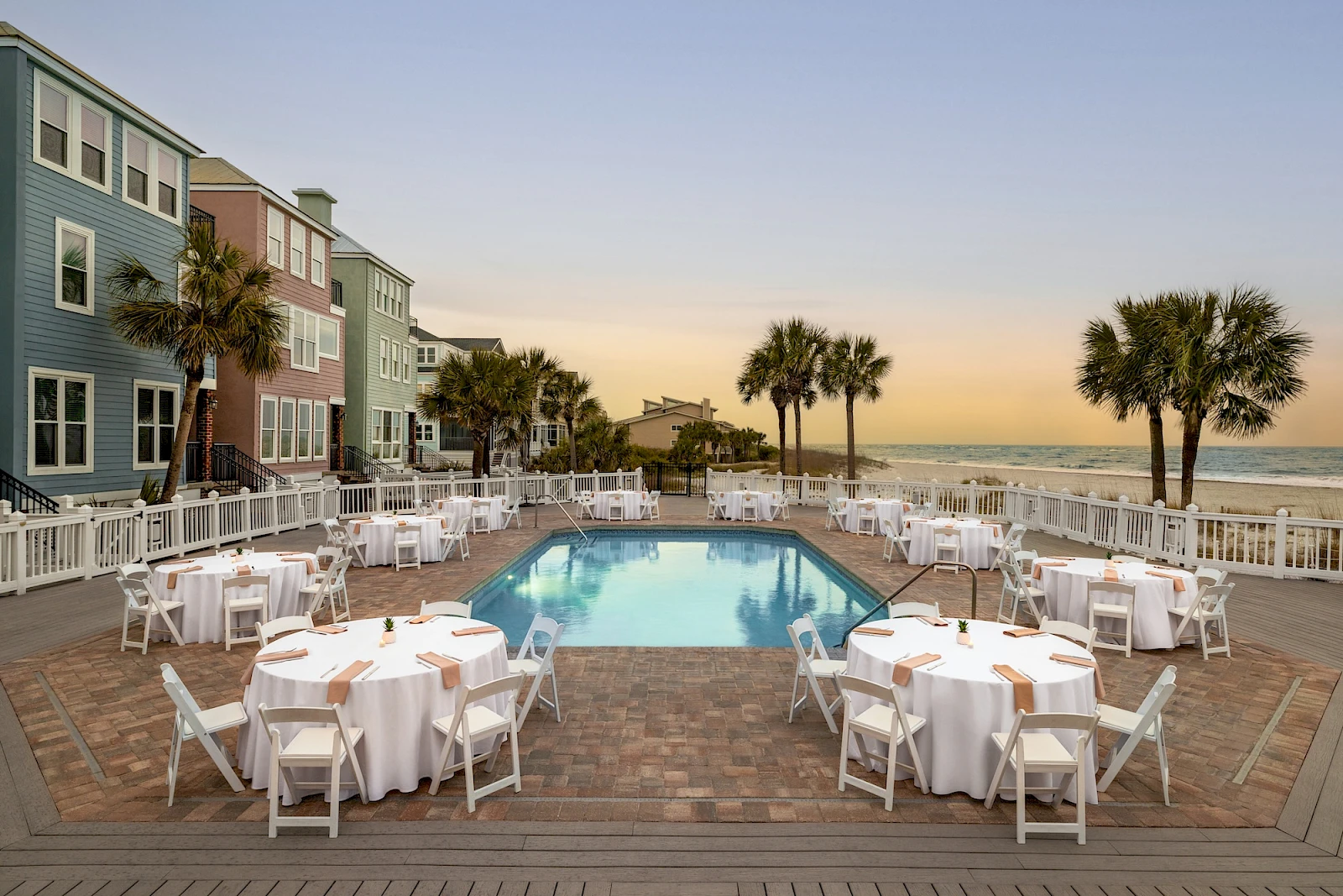 A resort pool area at sunset with round tables set for dining, white chairs, and palm trees near the ocean, overlooking the beach.