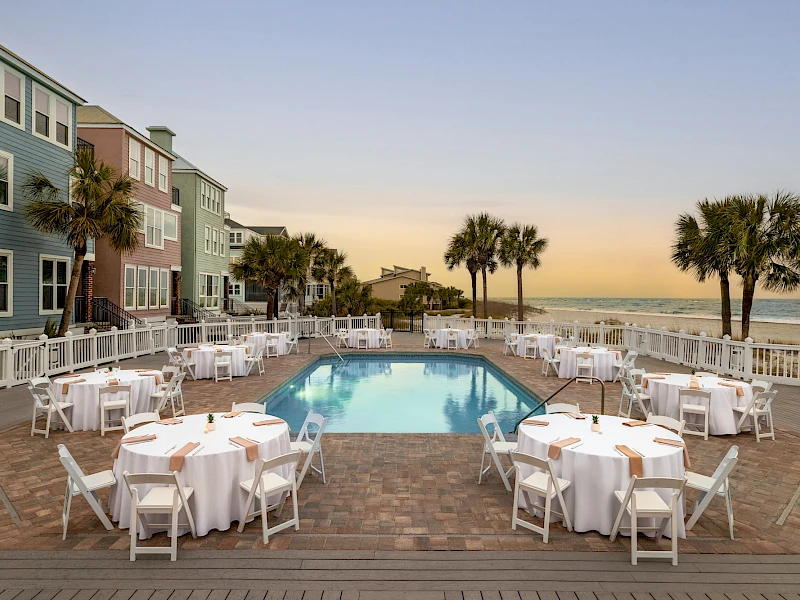 A resort pool area at sunset with round tables set for dining, white chairs, and palm trees near the ocean, overlooking the beach.