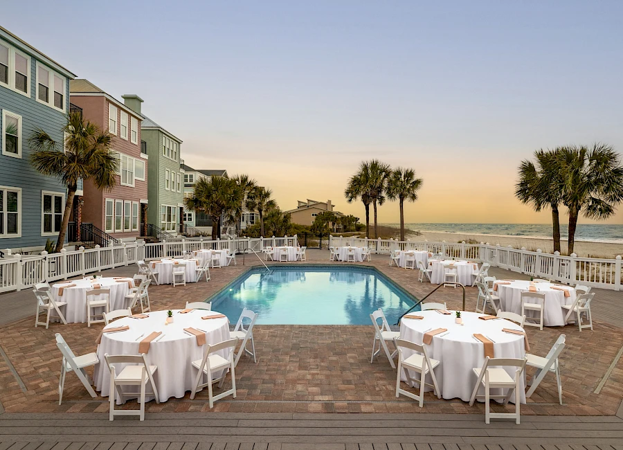 A resort pool area at sunset with round tables set for dining, white chairs, and palm trees near the ocean, overlooking the beach.