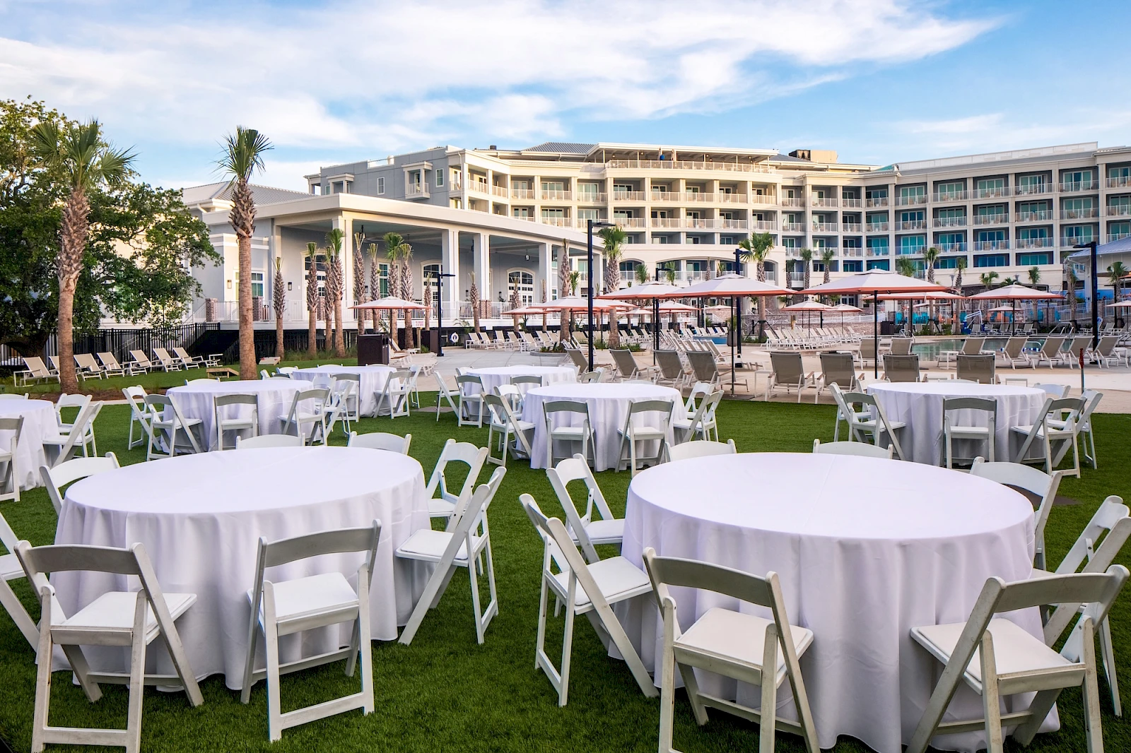 An outdoor event setup with round white-draped tables and white chairs on a grassy lawn near a modern hotel, ready for a reception.