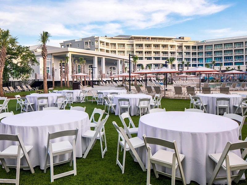 Outdoor hotel event setup with white round tables and folding chairs on a grassy lawn, a pool area, and a resort building in the background.