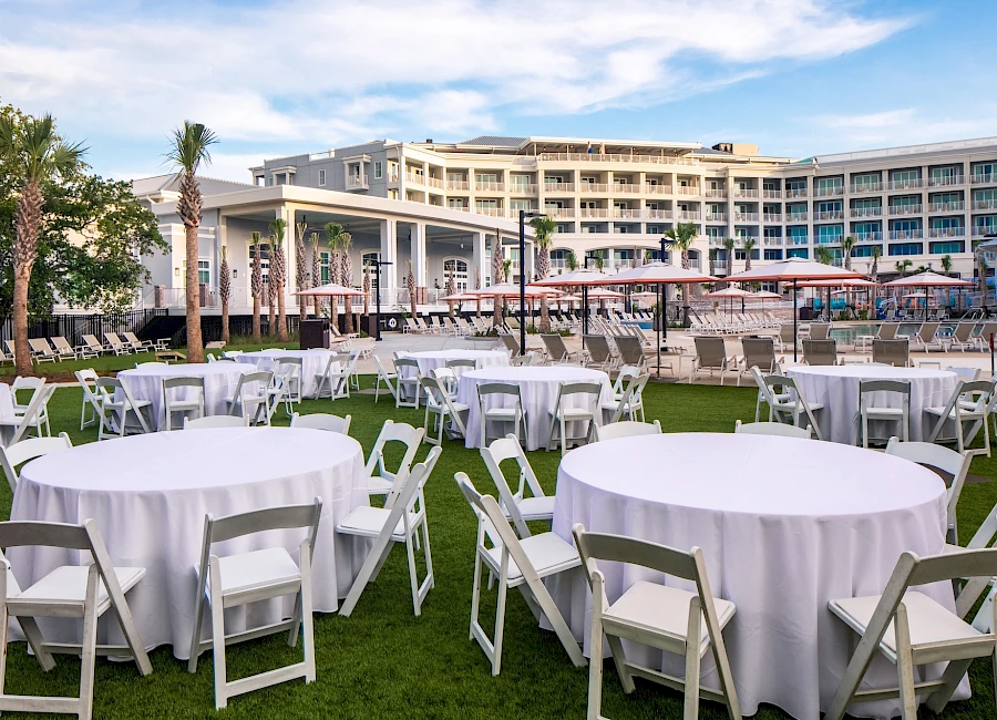 Outdoor hotel event setup with white round tables and folding chairs on a grassy lawn, a pool area, and a resort building in the background.