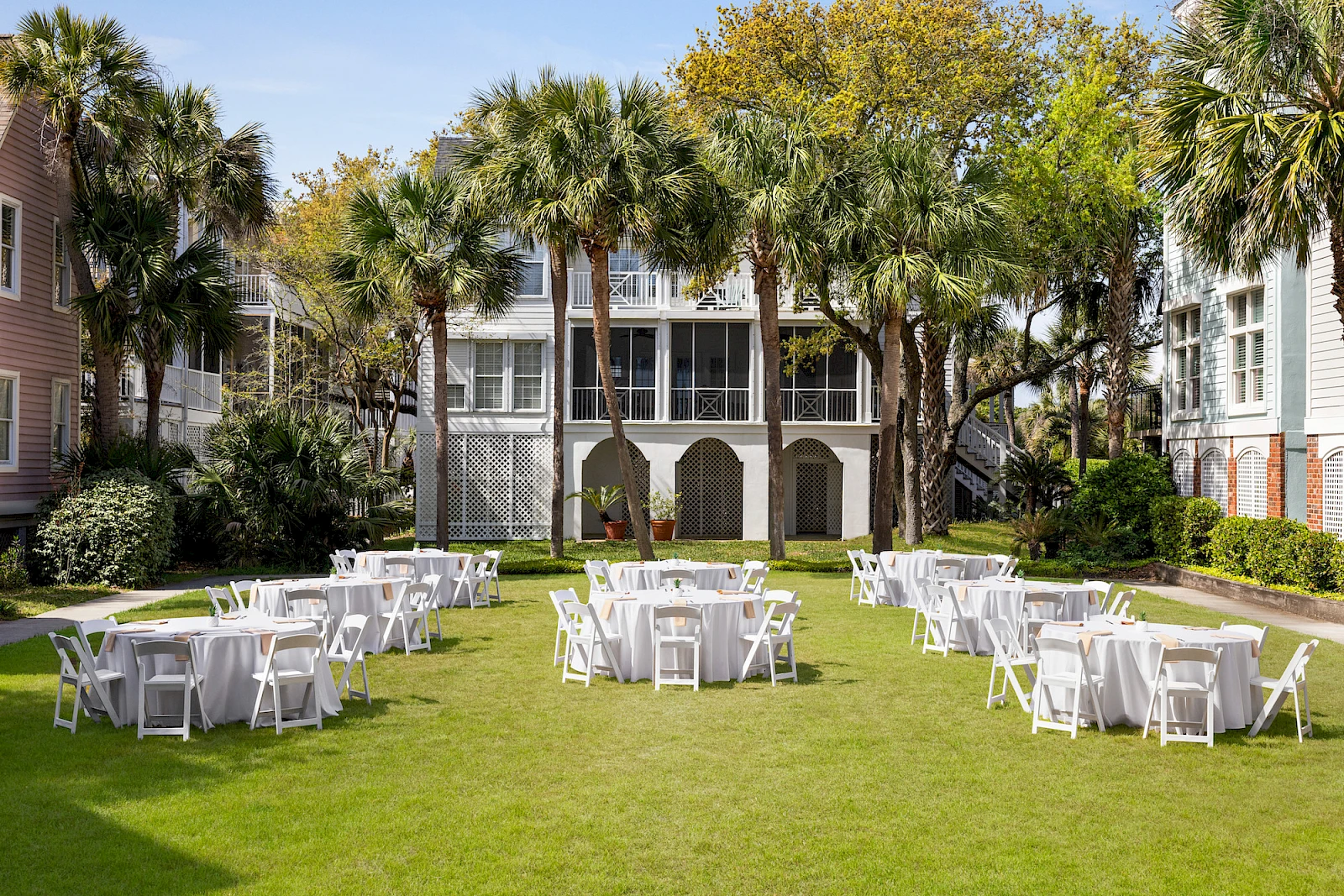 A sunny outdoor ceremony setup with white chairs and tables arranged on a green lawn, surrounded by palm trees and a charming building backdrop.