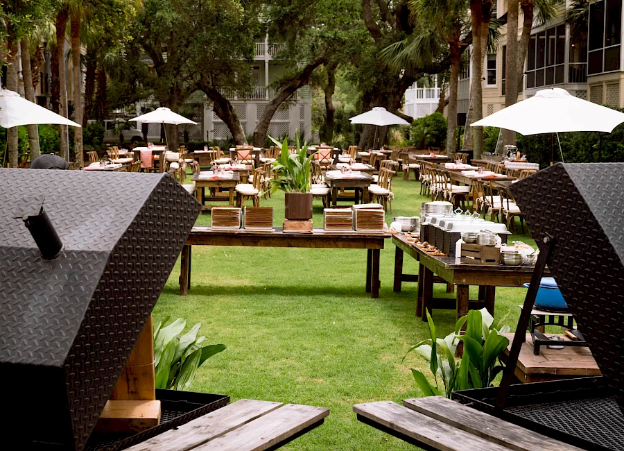 A daytime outdoor dining area with many wooden tables, chairs, umbrellas, and lush green lawn surrounded by trees.