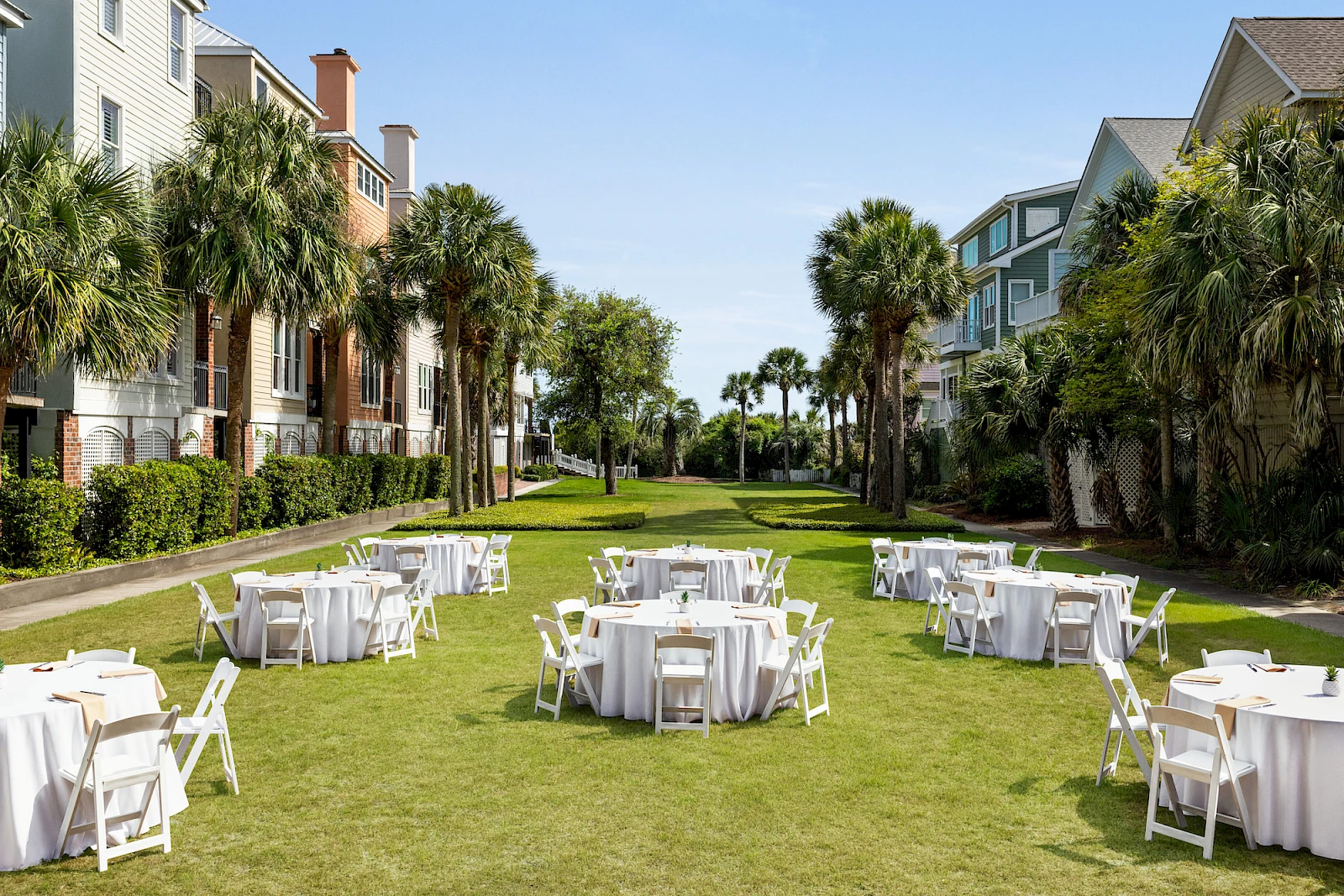 Outdoor party setup on a sunny lawn with round tables and white chairs, between resort-like buildings and palm trees, ready for guests.