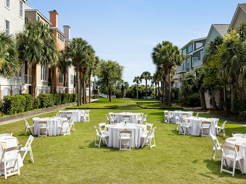 Outdoor party setup on a sunny lawn with round tables and white chairs, between resort-like buildings and palm trees, ready for guests.