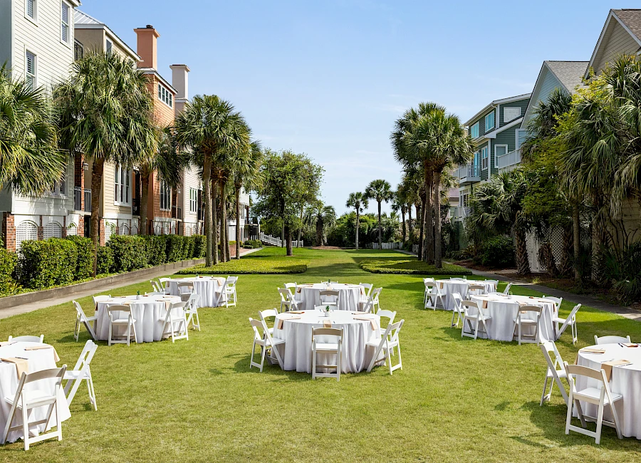 Outdoor party setup on a sunny lawn with round tables and white chairs, between resort-like buildings and palm trees, ready for guests.
