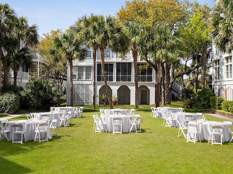Outdoor wedding or event setup on a green lawn with white folding chairs and round tables, surrounded by palm trees and a building backdrop.