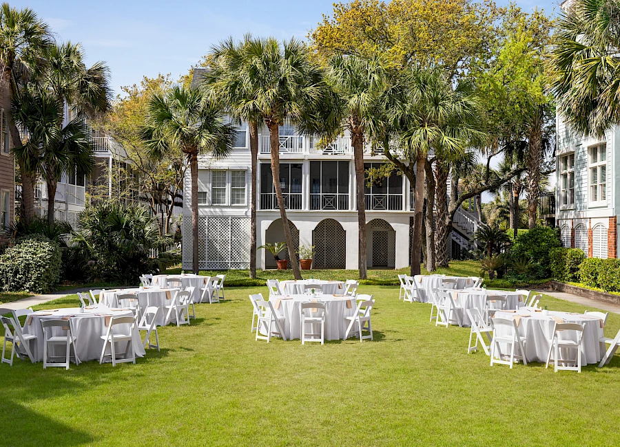 Outdoor wedding or event setup on a green lawn with white folding chairs and round tables, surrounded by palm trees and a building backdrop.