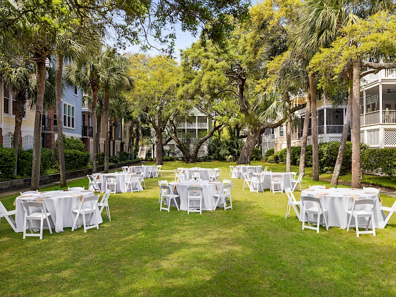 An outdoor event setup on a green lawn with round tables and white chairs arranged under trees, in a residential courtyard.