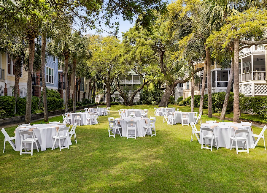 An outdoor event setup on a green lawn with round tables and white chairs arranged under trees, in a residential courtyard.
