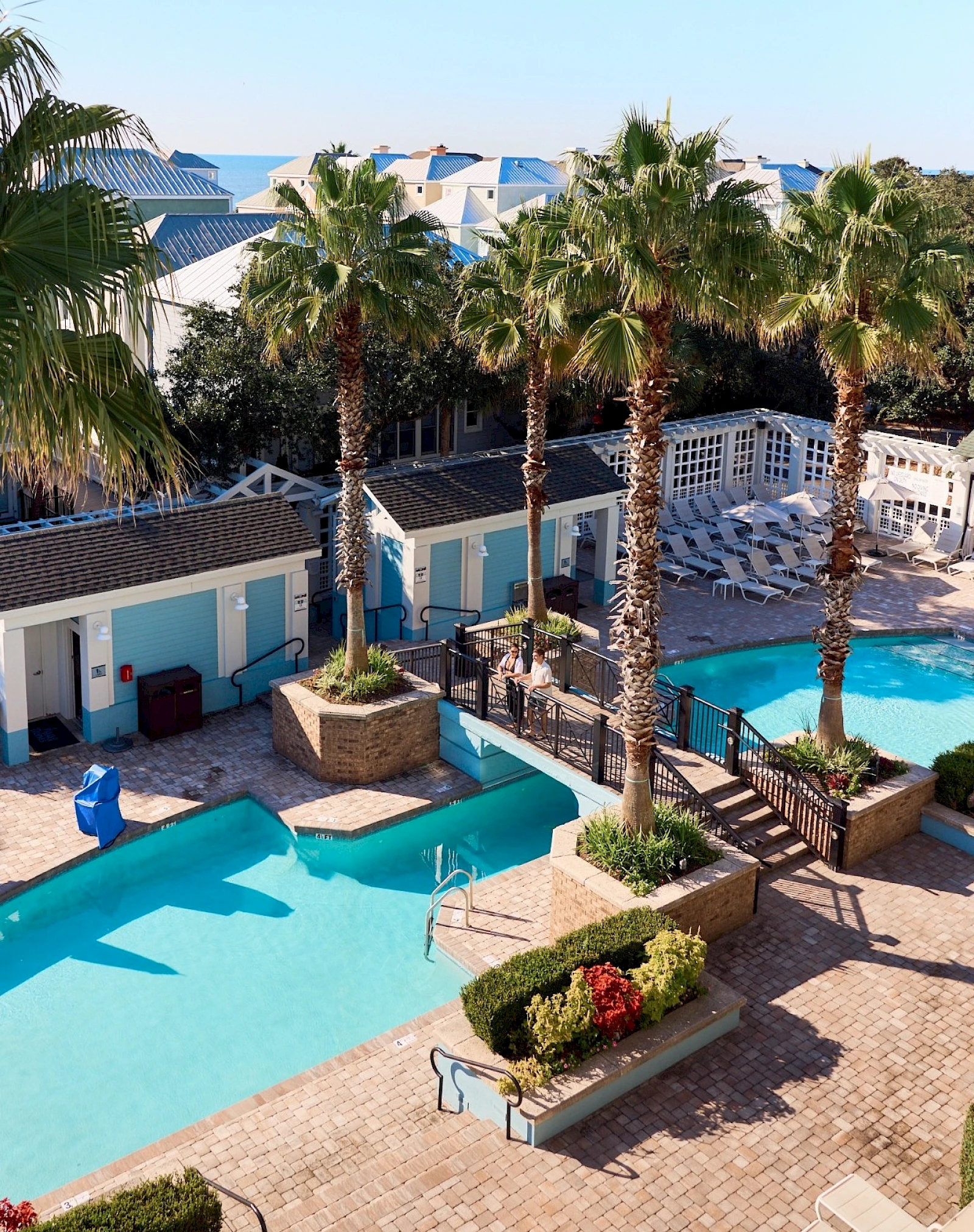 A resort pool area with turquoise pools, palm trees, sun loungers, white railings, and blue-roofed cabanas surrounding a sunny courtyard.