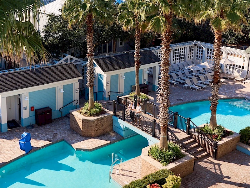 A resort pool area with turquoise pools, palm trees, sun loungers, white railings, and blue-roofed cabanas surrounding a sunny courtyard.