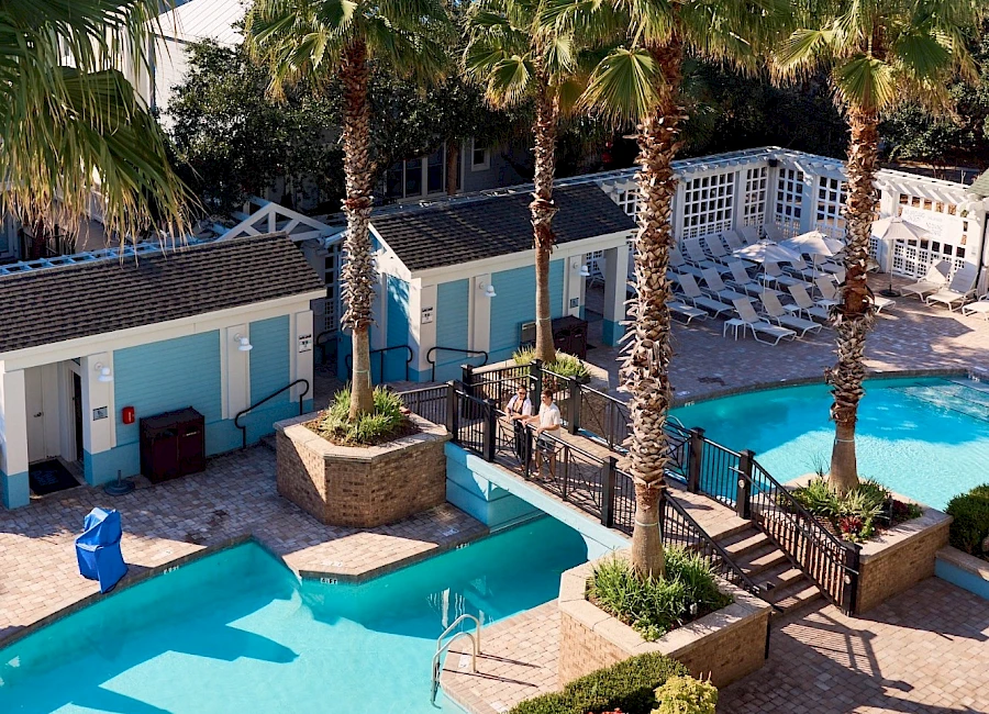 A resort pool area with turquoise pools, palm trees, sun loungers, white railings, and blue-roofed cabanas surrounding a sunny courtyard.