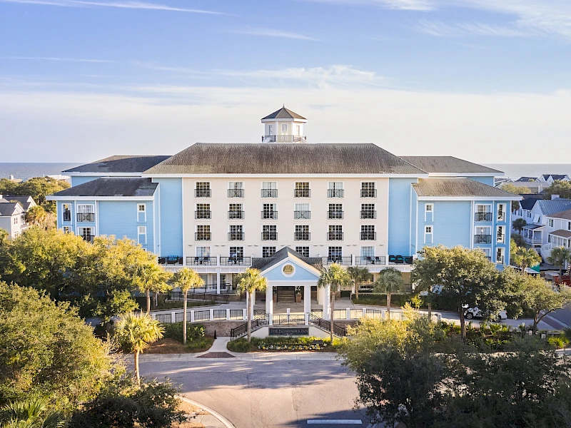 A light blue, four-story seaside hotel with a central portico, white-framed windows, palm trees lining the entrance, and a calm ocean view in the background.