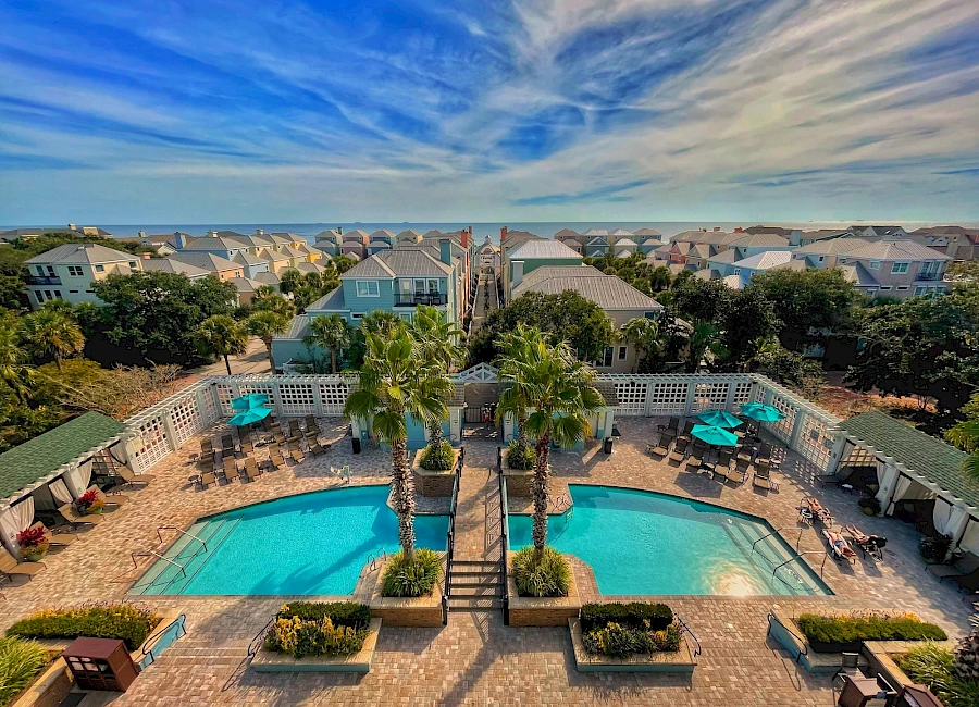 Aerial view of a sunny hotel pool area with two turquoise pools, palm trees, surrounding lounge chairs, and a breezy coastal neighborhood beyond.
