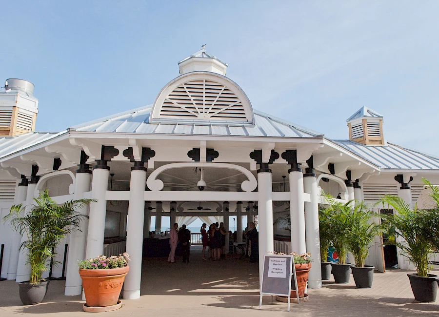 A sunlit, white colonial-style entrance with arched porch, columns, potted plants, and people inside, likely a venue or event hall.