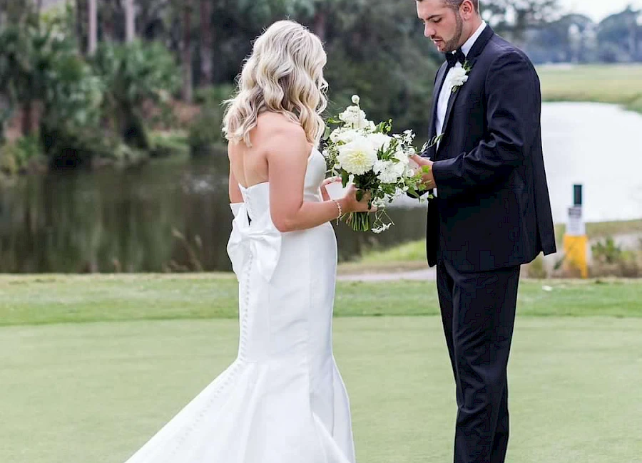 A newlywed couple stands on a lush lawn by a lake, the bride in a white gown holding a bouquet while the groom in a dark suit reads from a note.