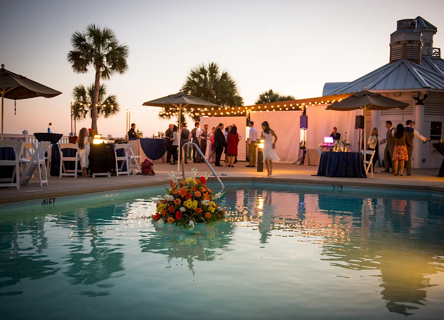 A poolside wedding reception at sunset: guests mingle near a lit terrace, flower centerpiece floating in water, palm trees, and a charming venue.