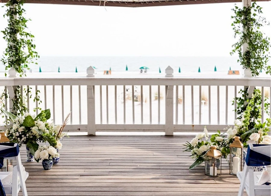 A beach wedding setup on a wooden deck with white chairs, floral arrangements, greenery, and a view of the sea, ending with a period.