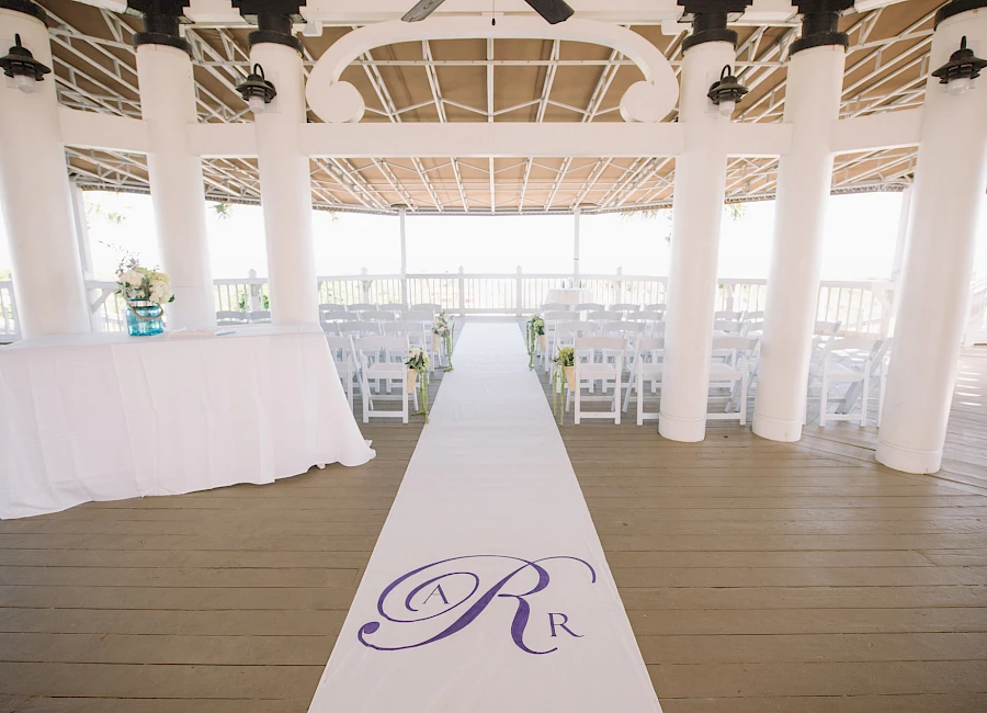 A bright wedding venue with a white aisle runner marked by a purple “R” logo, flanked by white chairs and a cake table inside a pavilion.