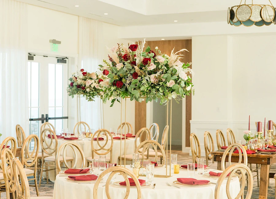 A bright dining area set for a formal event with round tables, floral centerpieces, and elegant wooden chairs. It’s airy and neatly arranged.