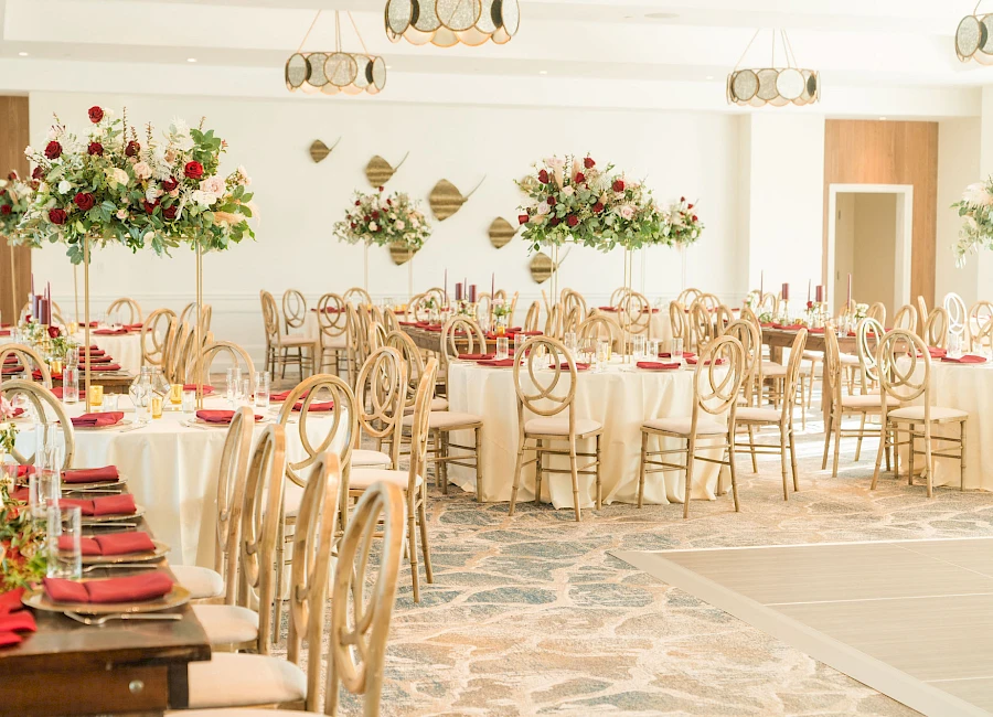 A bright event hall with white decor, long tables set with red napkins, elegant wooden chairs, and tall floral centerpieces in a festive reception setup.