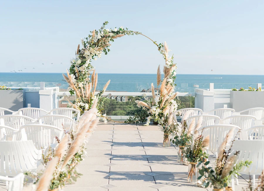 A seaside wedding aisle with a floral arch, white chairs lining a tiled path toward the ocean on a sunny day.