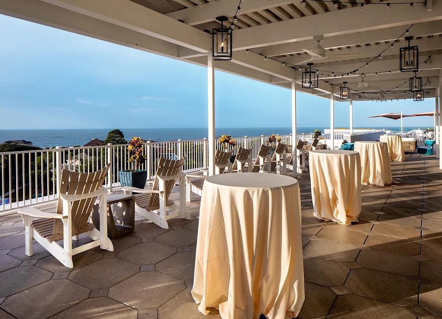 Ocean view terrace with round and high-top tables, beige tablecloths, white wooden chairs, and distant blue sea under a shaded pavilion.