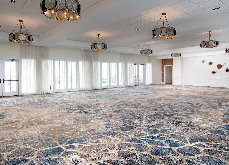 A large, empty room with stone-patterned carpet, numerous ceiling light fixtures, white walls, and big windows along one side.