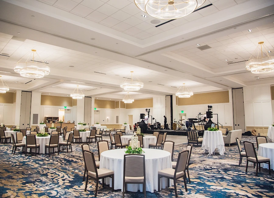 A large hotel banquet hall set with round tables draped in white cloths, potted greenery centerpieces, and many chairs arranged for a formal event.