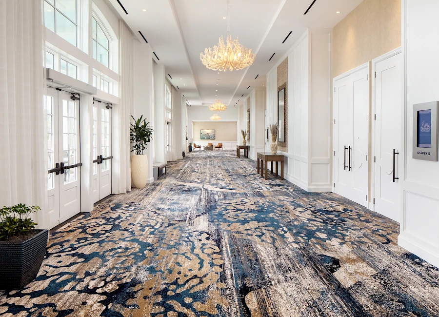 A luxurious hotel hallway with elegant chandeliers, white walls, long carpet, and potted plants along the sides, ending in a bright lobby.