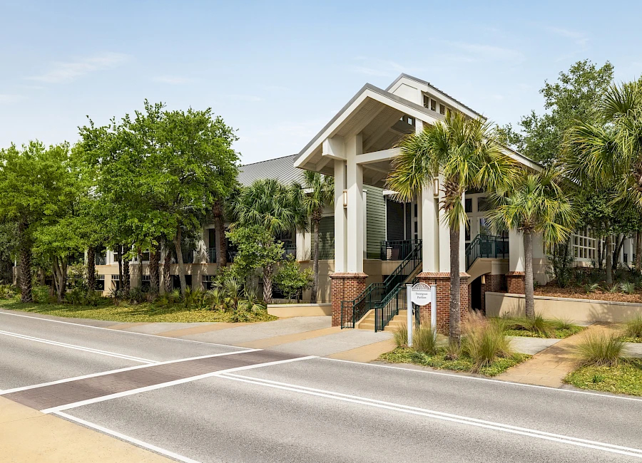 A low, modern building with a tall, peaked entrance, surrounded by palm trees and shrubs along a street, sunny and calm today.