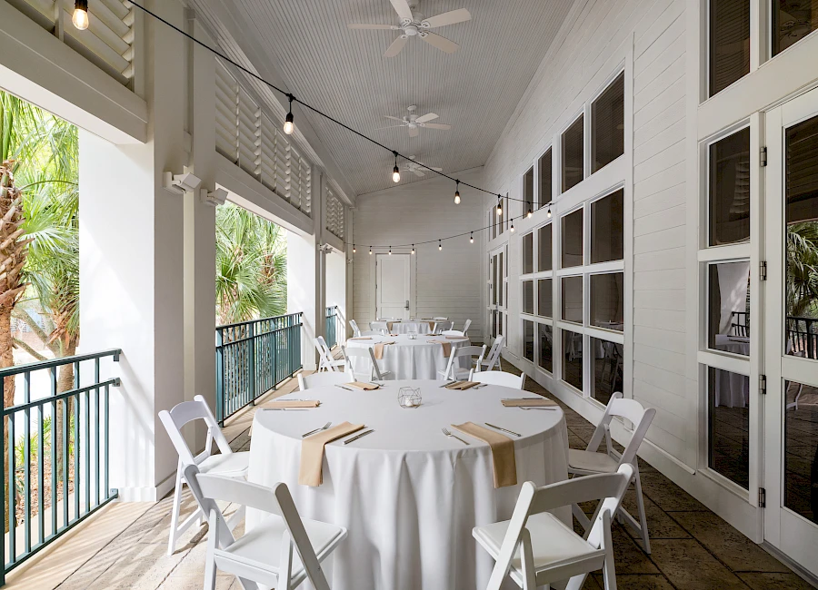 An airy porch setup with round tables, white chairs, white linens, and warm string lights; tropical plants outside and large windows to the side.