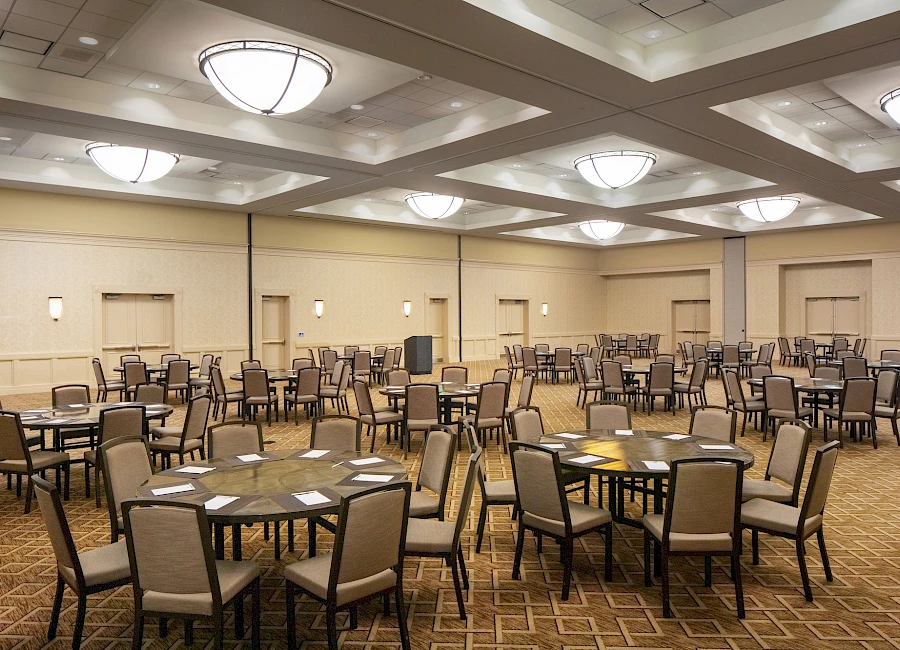 A large banquet hall set with multiple round tables, chairs arranged for an event, neutral walls and soft ceiling lighting.