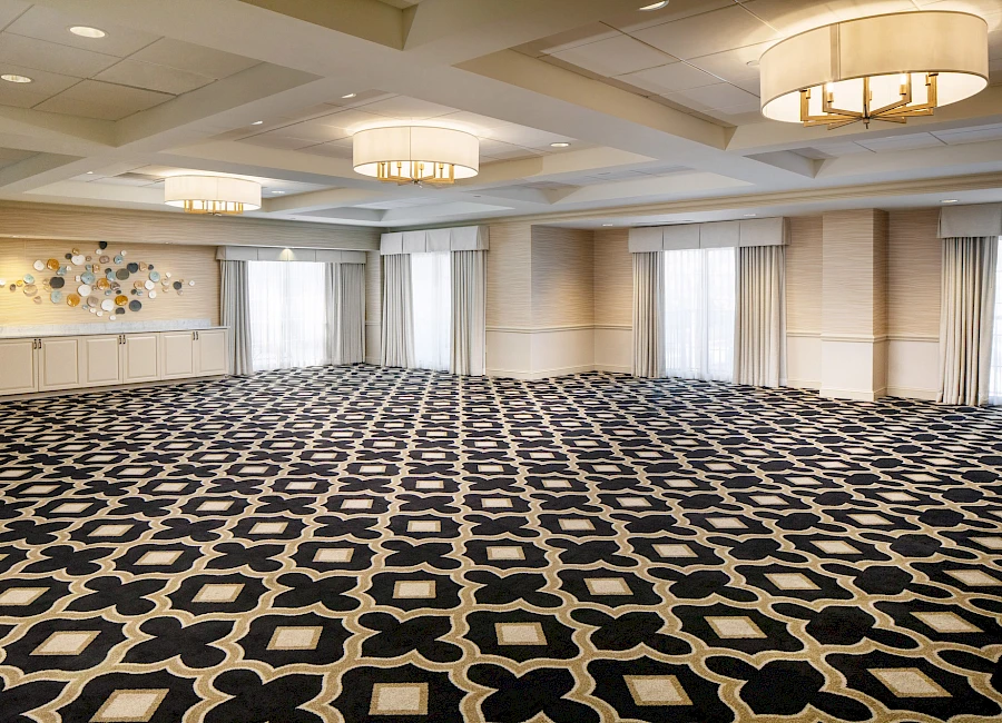 A large, empty conference hall with a black and white geometric carpet, round chandeliers, and long curtains along the windows, awaiting setup.