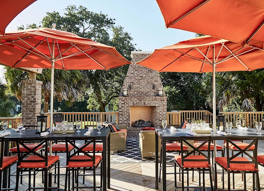 A sunny outdoor dining setup with brick grill, long tables, orange chairs, and large orange umbrellas around a central fireplace.