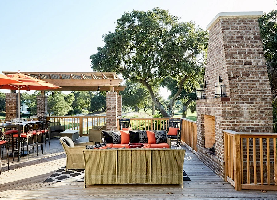 A sunny outdoor patio with wicker seating, orange umbrellas, a brick grill area, and a wooden railing overlooking trees, perfect for lounging.