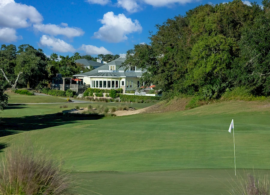 A lush golf course with a green, flagstick hole, and a large clubhouse in the background under a blue sky with fluffy clouds.