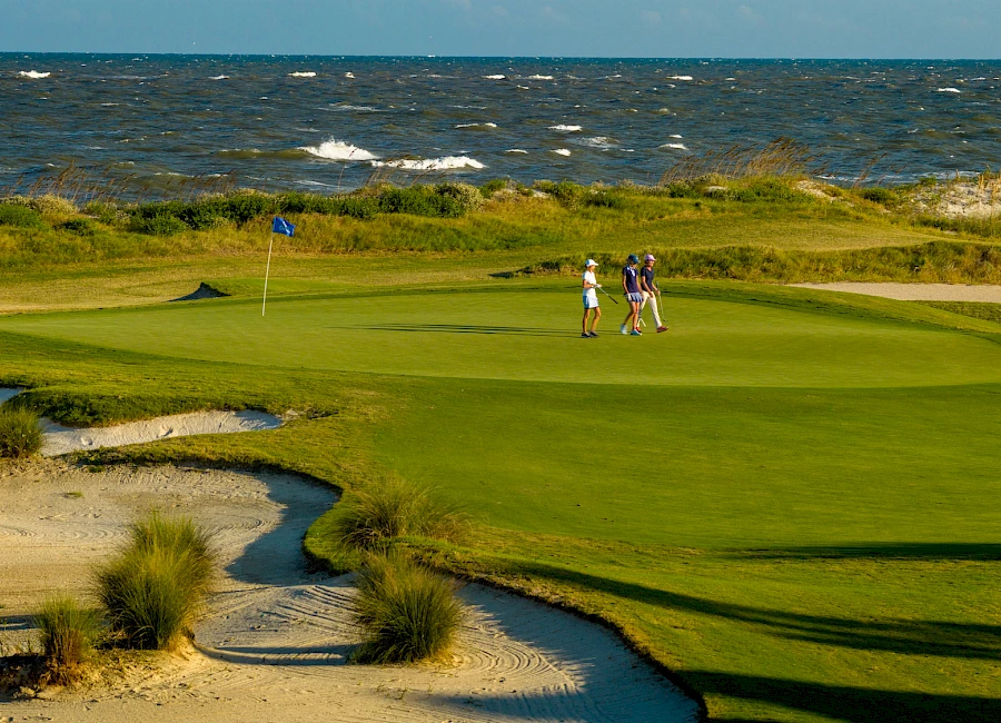 Two golfers walk on a sunlit coastal golf course, green fairways near sandy dunes, with the ocean in the background, under a bright sky.