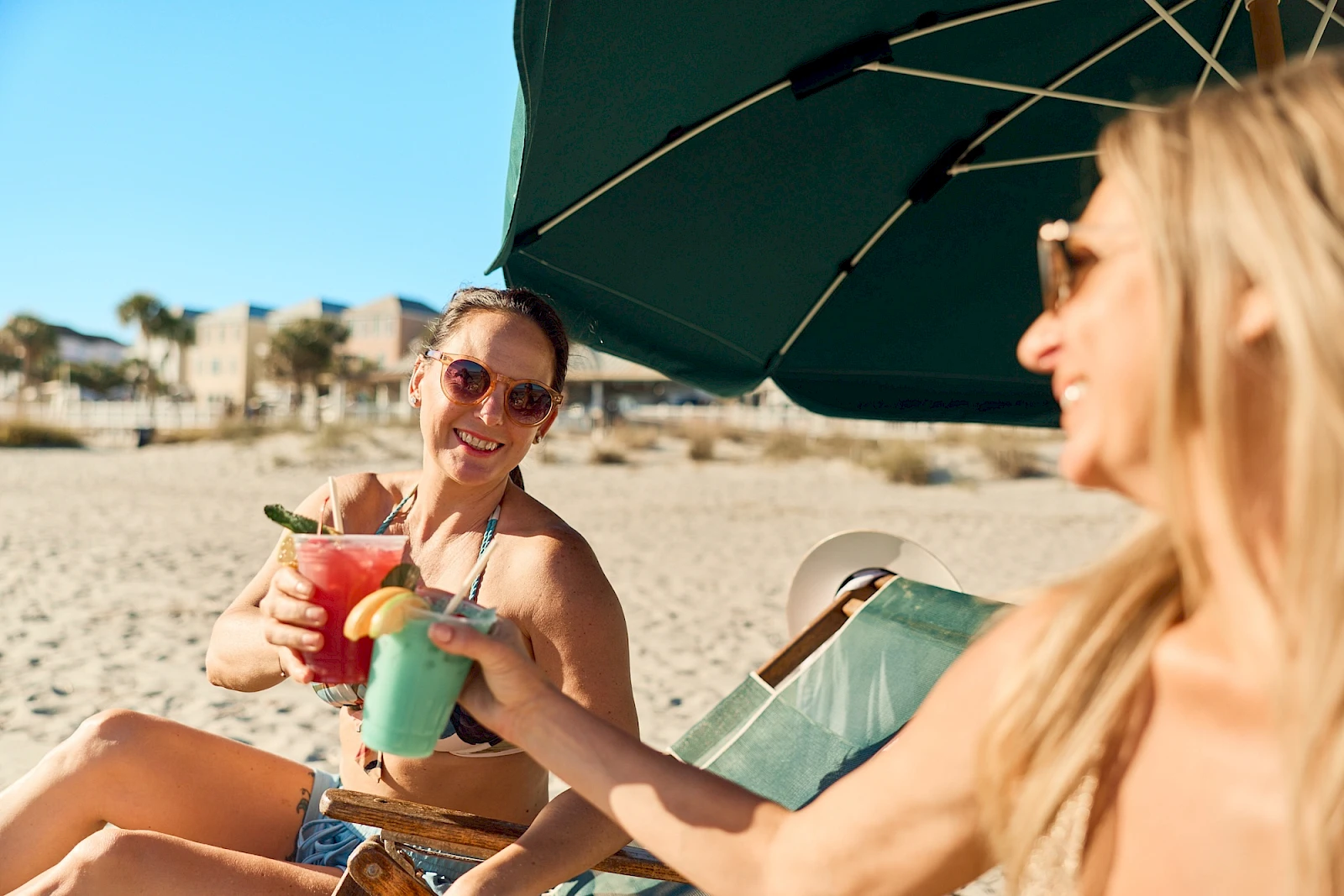 Two women on a sunny beach share colorful cocktails under a large umbrella, smiling and toasting with raised drinks.
