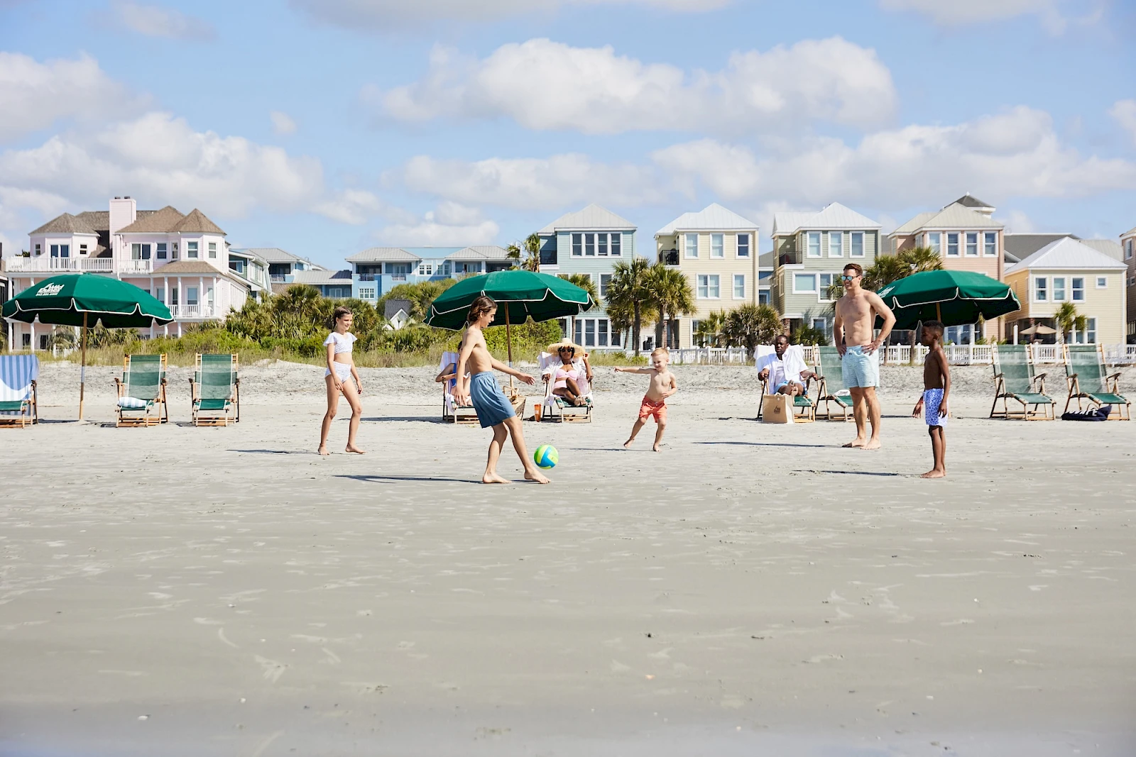 People playing on a sandy beach with colorful houses, umbrellas, and lounge chairs in the background, kids kicking a ball and adults watching.