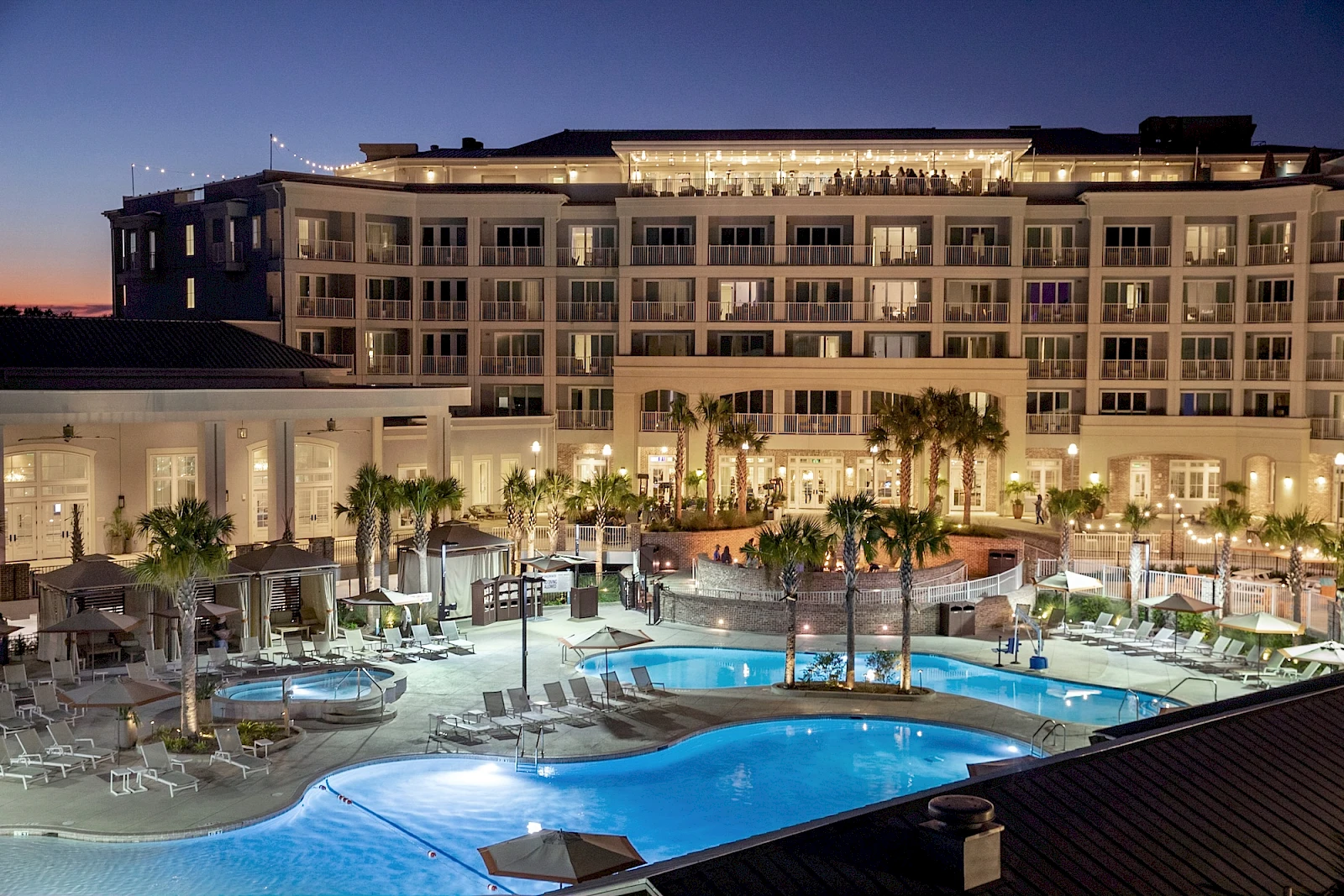 A luxury hotel with a large illuminated pool area, lounge chairs, palm trees, and a multi-story building at dusk, bustling with guests.