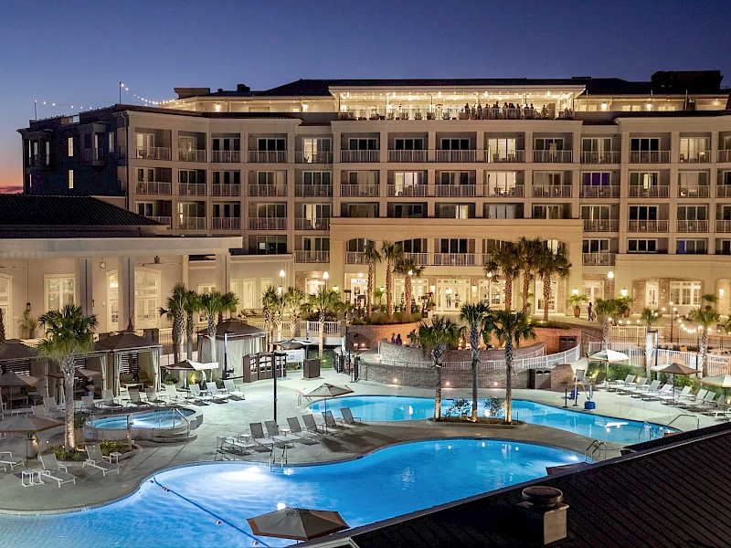 A luxury hotel with a large illuminated pool area, lounge chairs, palm trees, and a multi-story building at dusk, bustling with guests.