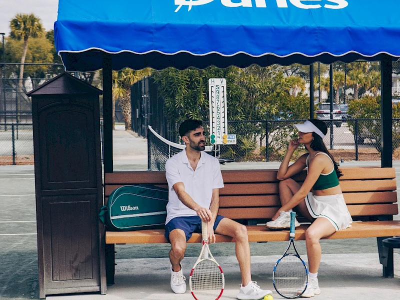 Two people sit on a bench under a blue “Wild Dunes” canopy at a tennis court, with rackets and a ball between them.