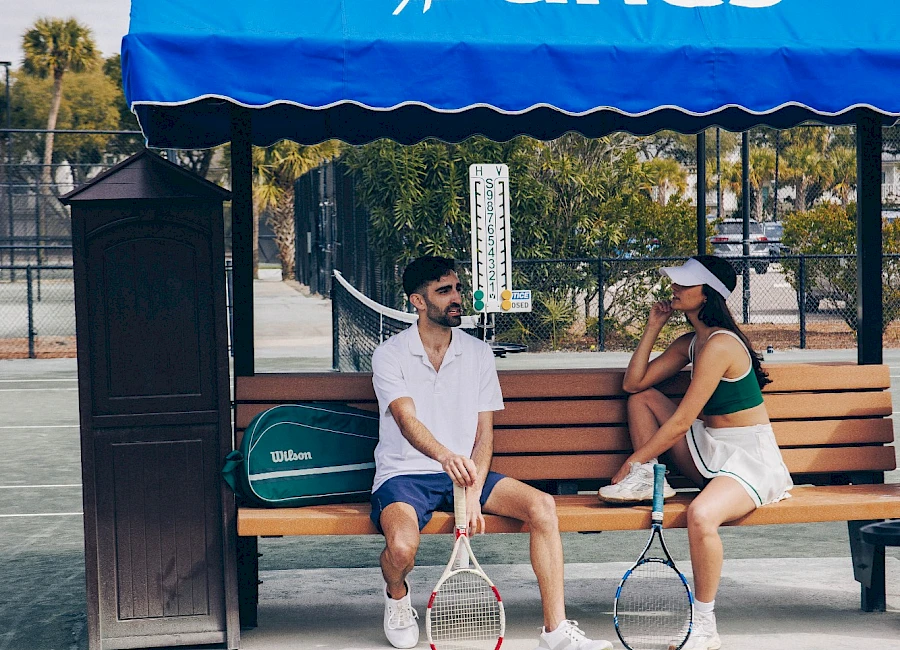 Two people sit on a bench under a blue “Wild Dunes” canopy at a tennis court, with rackets and a ball between them.