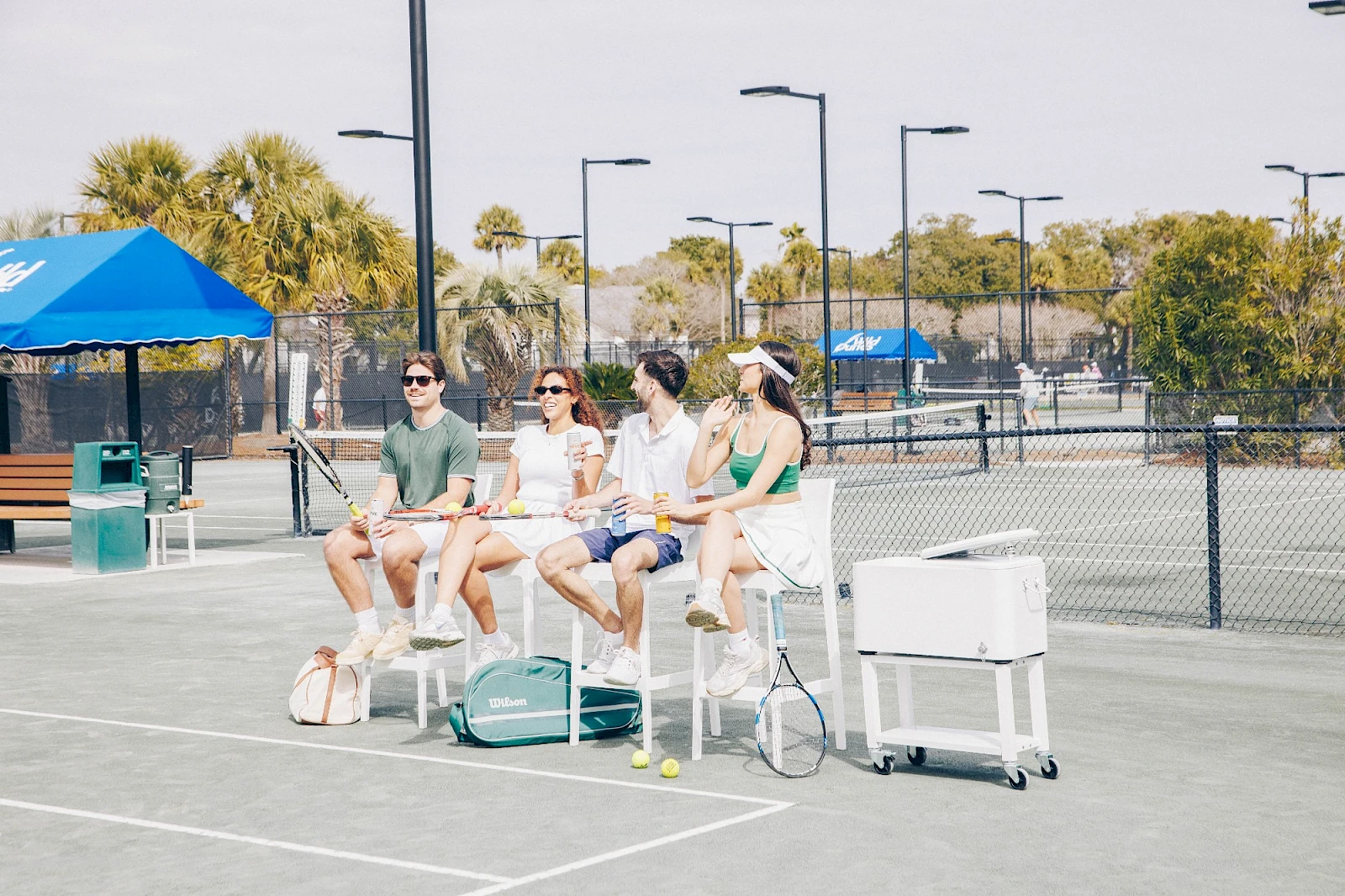 Four people sit on tall chairs at a sunny outdoor tennis court, with bags and a cooler nearby, watching something off-camera.